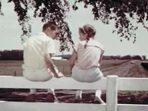 A boy and a girl sitting on a white wooden fence outdoors.