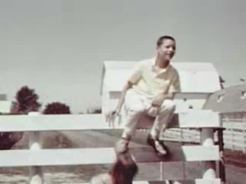 A young boy sitting on a white wooden fence waving his hand.
