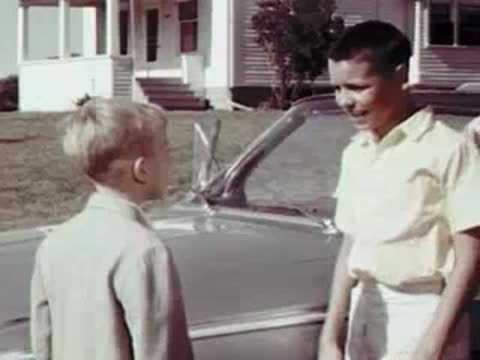 A young boy greeting another boy standing next to a parked car.