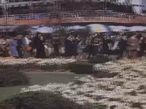 Pan right over crowds walking past flowerbeds outside exhibition halls at the World's Fair.