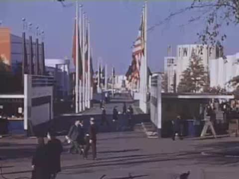 Pan right down a boulevard lined with flags and modern exposition buildings.