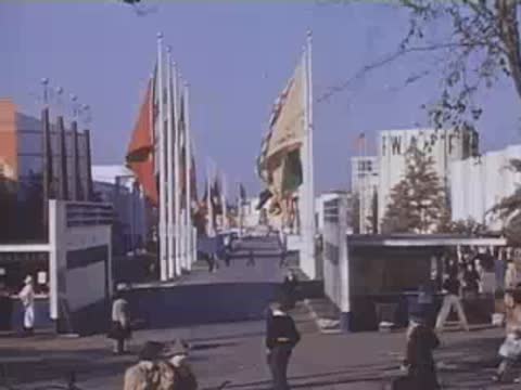 Pan right along a pathway flanked by tall flagpoles and exhibition pavilions with crowds.
