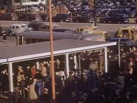 Pan right over a crowded parking lot filled with 1930s automobiles and buses.