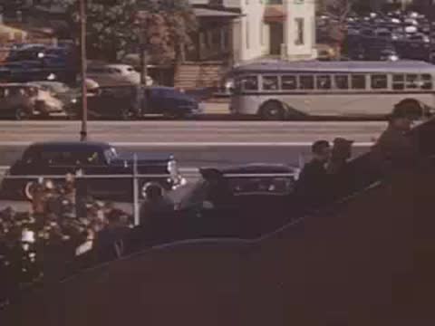 Pan right across a busy street scene featuring vintage cars and spectators watching.