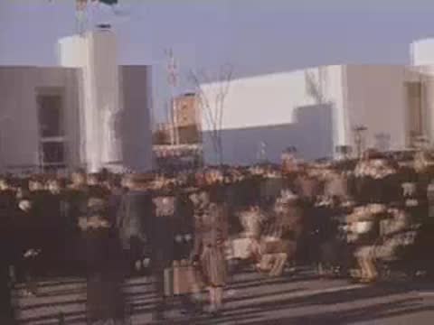 Pan right across crowds walking in front of white pavilions beneath fluttering flags.
