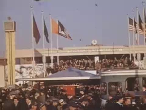 Pan right past a mural showing workers with flags and crowds gathered below a grandstand.