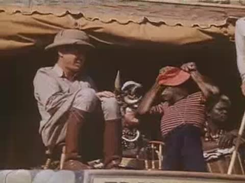 A chimpanzee adjusts a red hat on its head near a seated safari guide.