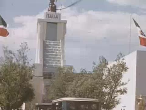 A medium shot of the ITALIA pavilion with flags flying and people walking by.