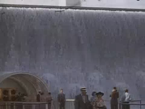 Water cascading down a large blue wall next to a public walkway