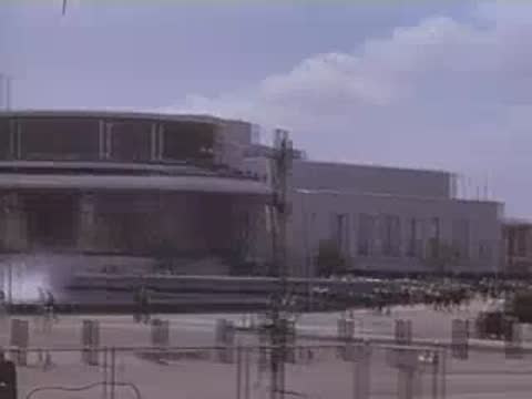 Wide shot of a modern pavilion building with water fountains and people walking nearby.
