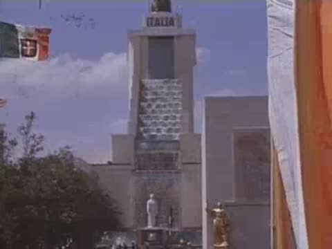 Wide shot of the Italy pavilion with flags and a vast parking lot filled with cars.