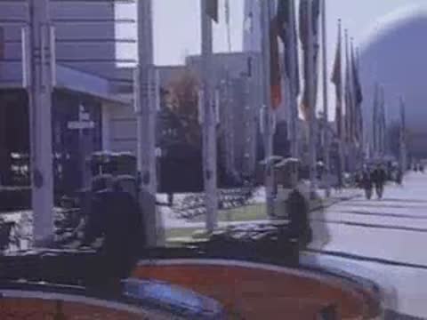 Panning shot moving from a packed parking lot towards a paved walkway with flags and buildings.