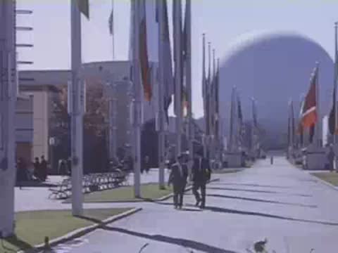 Walking down a paved boulevard lined with international flags toward a large white spherical pavilion.