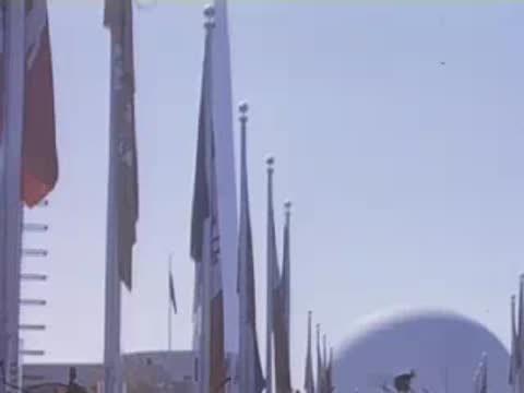 Low-angle shot looking up at tall flagpoles flying various national flags against a bright blue sky.