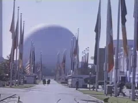 People walking on a wide paved path towards the Trylon and Perisphere structures with surrounding fountains.