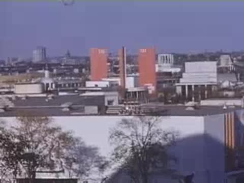 An elevated panoramic view over modern white exhibition buildings with bright orange brick towers in the distance.