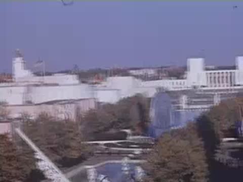 An elevated shot looking down at exhibition structures, a reflecting pool, and visitors walking on pathways.