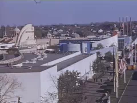 An elevated shot capturing modern exhibition buildings, flags, and visitors walking along a walkway.
