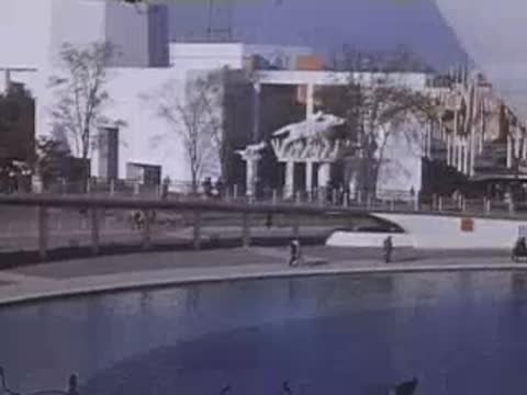 Looking across a reflecting pool toward an exhibition building featuring prominent sculptural statues on its roof.
