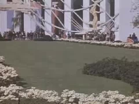 Pan right across grass toward buildings with crowds under white lattice archways at the World's Fair.