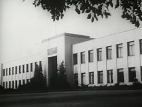 Exterior of a large factory building with trees in the foreground, 1940s film.