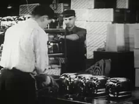 Two factory workers working near stacked products and toasters on a line, historical footage.