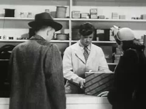 Customers and a store clerk at a retail shop counter looking at merchandise, 1940s.