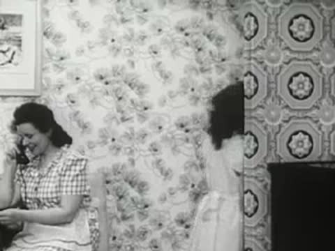 Woman talking on a telephone while other family members move around in the room, 1940s.