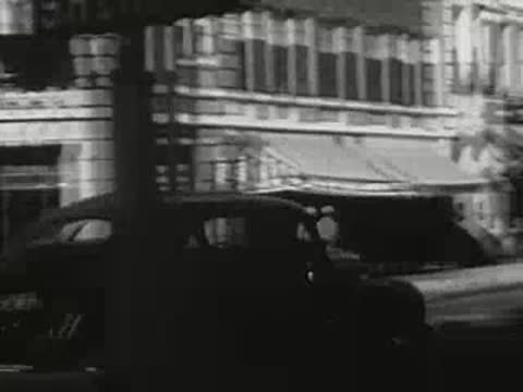 Car driving along downtown street with storefronts, 1940s.