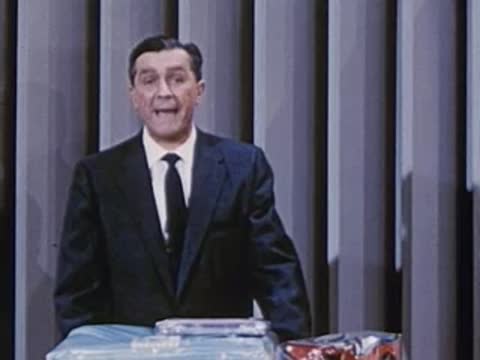 man in dark suit standing behind table displaying packaged items