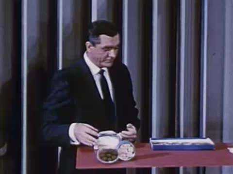 Man in suit stands behind desk with cans and packaged items