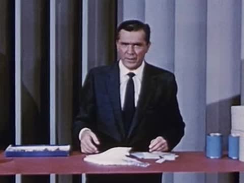 Man in suit stands behind desk pointing at folded white material