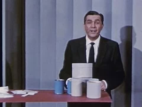 Man in suit stands behind desk with stacked cylindrical containers and cans