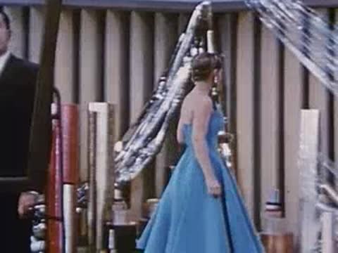 a woman in a blue gown walks past a man and shiny materials