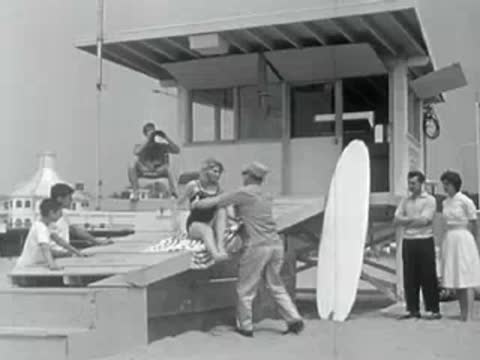 Lifeguards and people gathering near a beach lifeguard tower with a surfboard in the foreground.