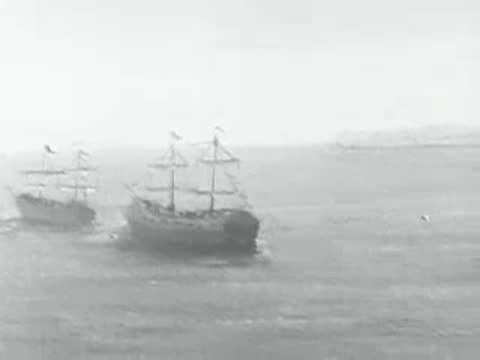 Historical sailing ships navigate rough ocean waters near rocks.