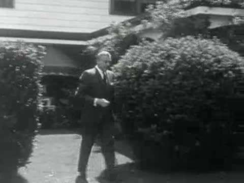 A man in a suit walks through a 1950s garden pathway.