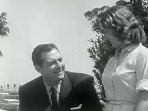 A man in a suit and a woman talk outdoors in 1950s California.