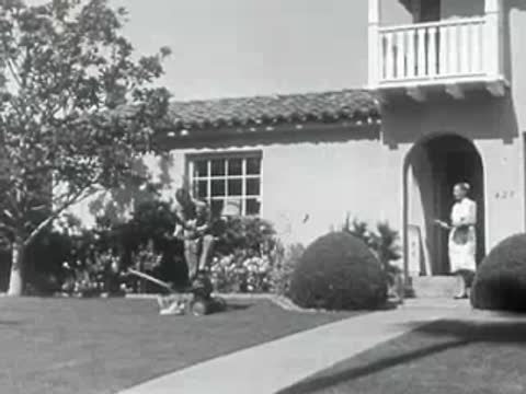 A man pushes a lawnmower in the yard of a house.