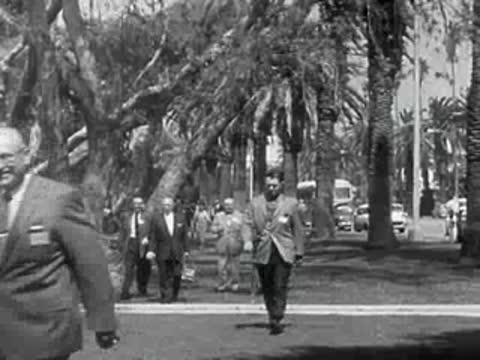 Men in suits walk through a park with palm trees in 1950s California.