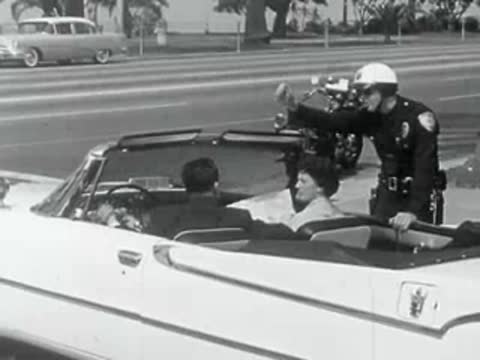 A police officer talks to people in a convertible car in 1950s California.