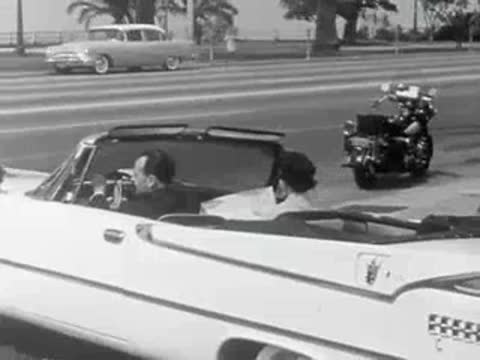 A white convertible drives along a coastal road with palm trees in 1959.