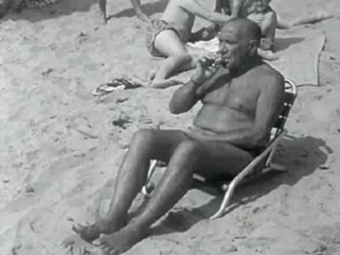 A woman sits on a beach towel while a man sunbathes in a chair.