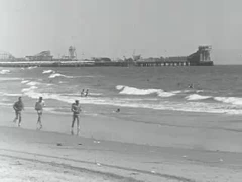 People run along the shoreline of a sandy beach near a pier.