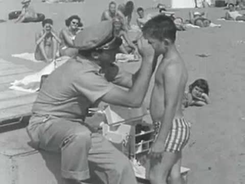 A lifeguard puts sunscreen on a young boy sitting on a box.