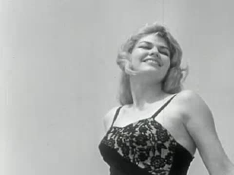 A close-up of a smiling woman posing in a black patterned swimsuit against a bright background.