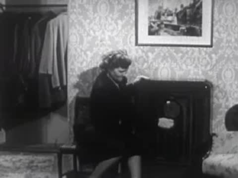 Woman standing indoors near a dark stove/fireplace in a room with patterned wallpaper.