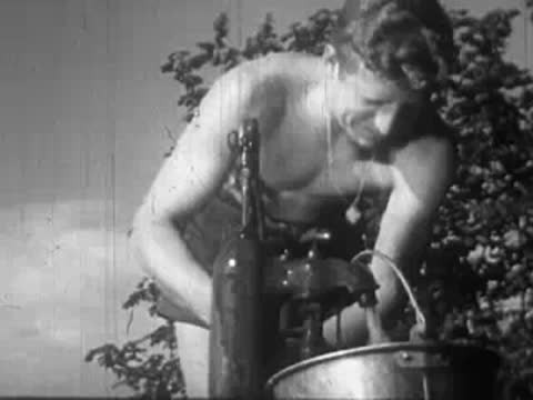 Shirtless man working with a rustic outdoor water pump or washing apparatus.