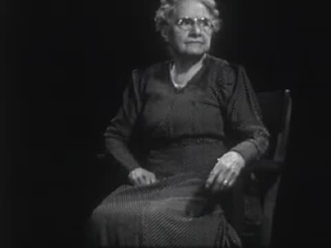 Elderly woman wearing glasses seated in a chair against a dark background.