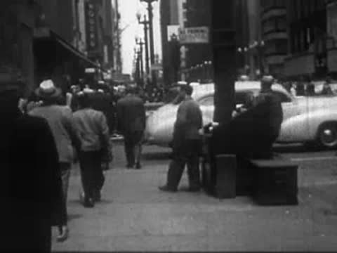Busy city street scene with pedestrians walking on sidewalk and cars passing.